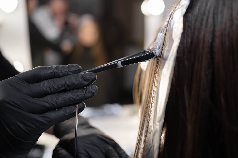 close up side view of a strand of female hair on a special sheet of foil, the master applies hair dye with a brush. The process of highlighting by a professional master in a beauty salon.